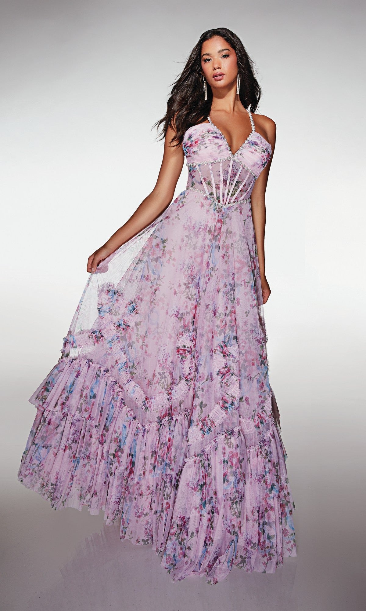 A woman stands against a plain background in the Alyce 62028 long prom dress, featuring a flowing floral print, sheer corset bodice, and ruffled hem.
